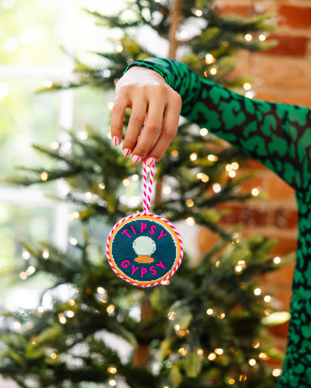 Person holding a 'Tipsy Gypsy' ornament in front of a decorated Christmas tree.