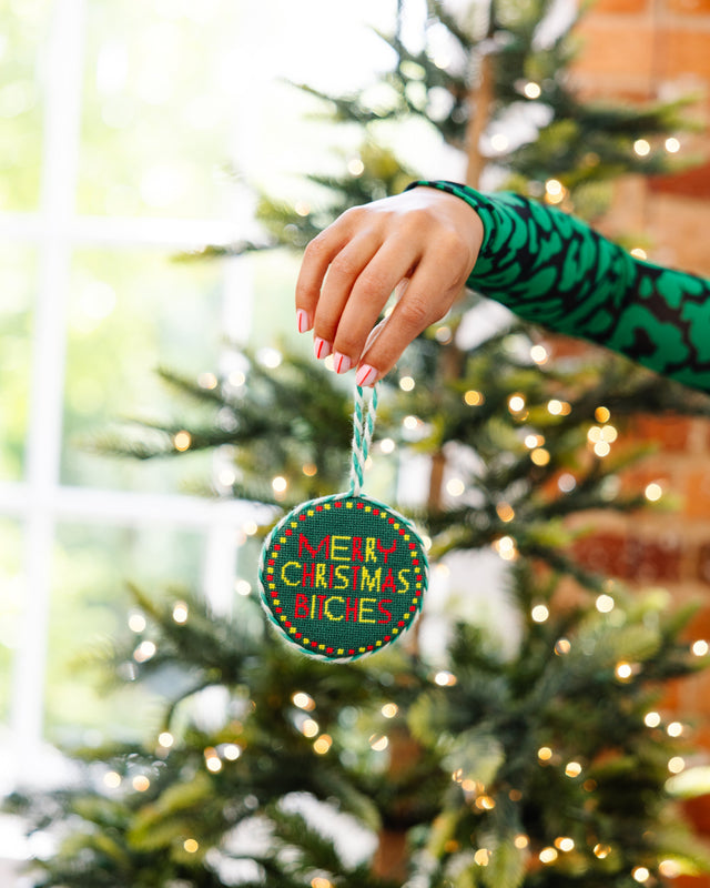 Hand holding a 'Merry Christmas Bitch' ornament in front of a decorated Christmas tree.