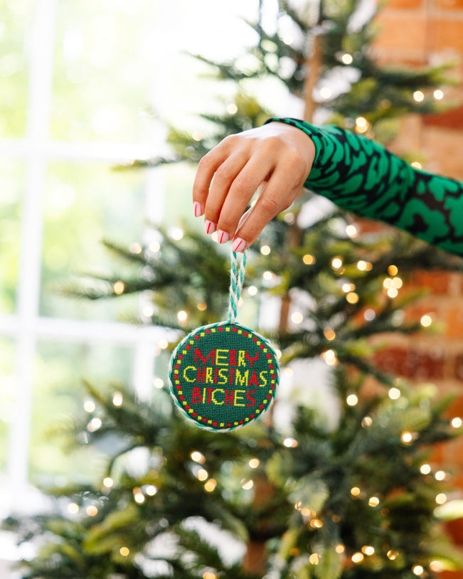 Hand holding a 'Merry Christmas Bitch' ornament in front of a decorated Christmas tree.