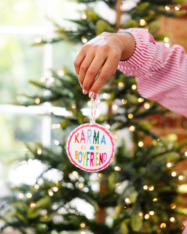 Hand holding a Christmas ornament with 'Karma is my boyfriend' text in front of a decorated tree.
