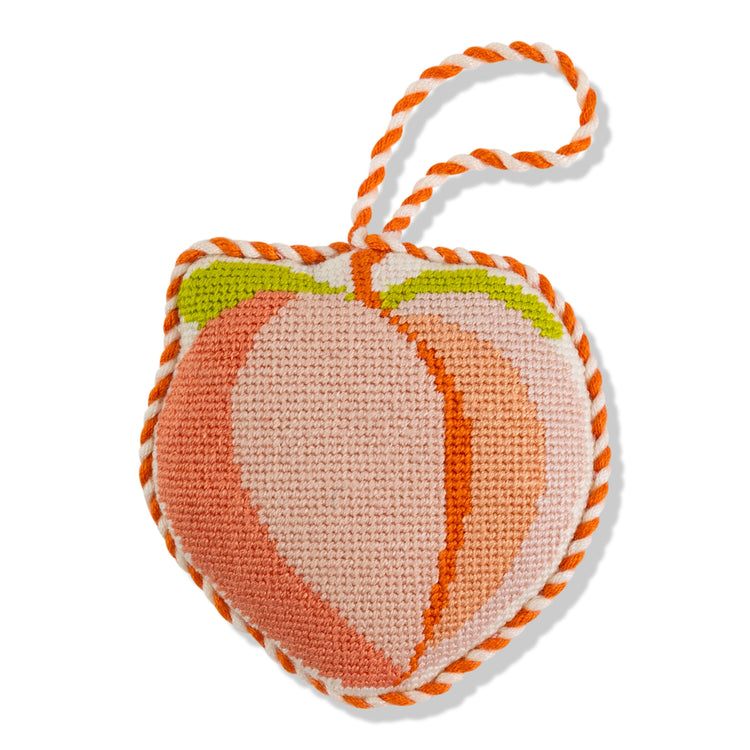 A needlepoint ornament in the shape of a peach emoji, with orange, green, and pink colors, backed with velvet and tied with a white and orange string.