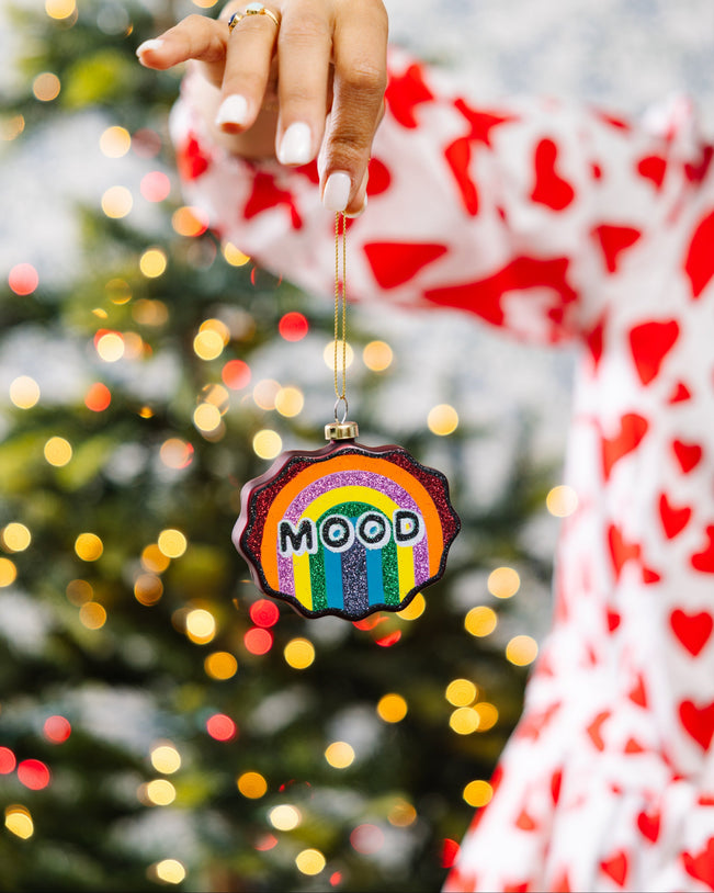 Hand holding a rainbow-colored ornament with 'MOOD' text against a Christmas tree background.
