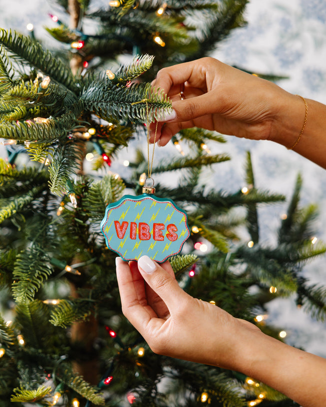 Hand holding a Christmas ornament with 'VIBES' text against a decorated tree background