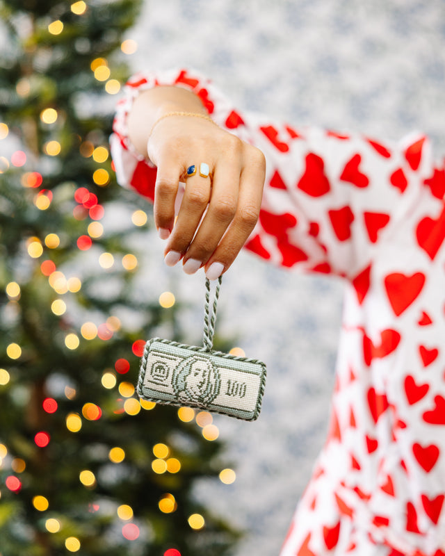 Hand holding a keychain with a dollar bill design in front of a blurred Christmas tree.