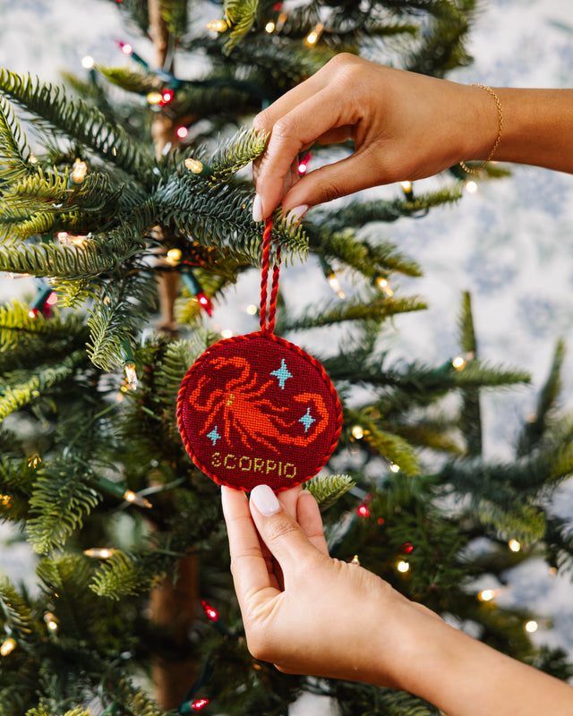 Hand holding a red 'Scorpio' ornament against a decorated Christmas tree.