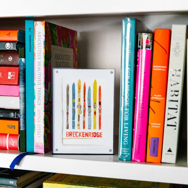 Breckenridge Matchbook - Furbish Studio, A Breckenridge Matchbook watercolor print featuring multiple colorful Ski boards arranged vertically enclosed in a 5" x 7" black magnetic acrylic floating frame sitting on a bookshelf in the middle of books